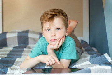 Naklejka premium a boy with blond hair in a blue t shirt is lying with a computer tablet and looks seriously at the camera