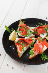 Sandwich with salmon, cheese, arugula and lemon on a black plate on a white table