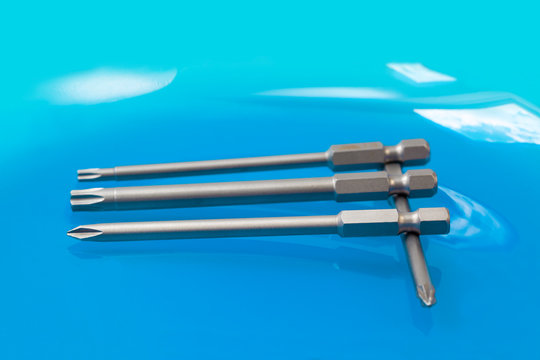 Three parallel arranged screwdriver bits for slotted-head and cross-head screws and positioned in a right angle to one cross-head screwdriver bit on turquoise background
