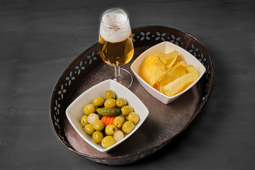 Closeup of olives with pickles, mini onions, carrot and chips in a white bowl, a beer in a crystal glass on a metallic bowl. Grey  wood background. Home or bar appetizer.