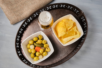 Closeup of olives with pickles, mini onions, carrot and chips in a white bowl, a beer in a crystal glass on a metallic bowl and a brown cloth. Grey  wood background. Home or bar appetizer.
