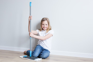 Girl 10 years old sitting on the floor with a mop. Mom’s assistant. Smiling. House cleaning. Grey...