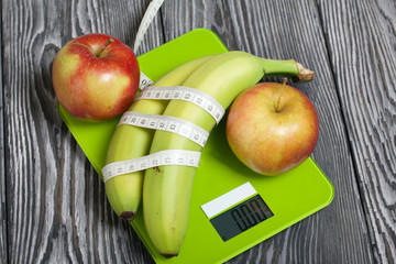 Bananas wrapped in measuring tape lie on the scales. Near apples. Scales show zero. On brushed pine boards. Symbol of World No Diet Day.