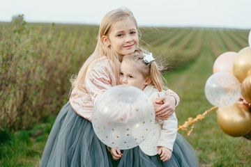 Two little sisters in luxurious dresses with balloons on a background of green field. daughter's day