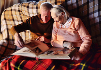two elderly people-a man and a woman looking at a family photo album smile and hug each other wrapped in a warm plaid blanket.