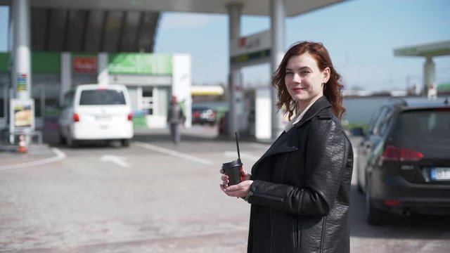 customer service, a happy young woman with a cup of coffee in her hands stands background of gas station while car is being fueled, petrol prices concept