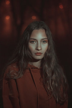 Portrait of a girl with long hair in a hoodie surrounded by darkness and red lights at night