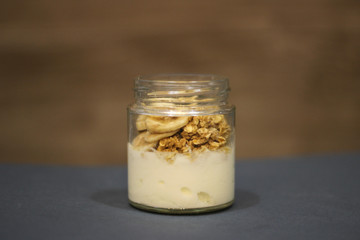 glass of yogurt with granola and banana on blue table and wooden background