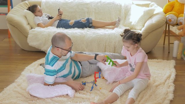 The father and daughter play together on the floor with the constructor, while the eldest daughter lies on the sofa and uses a smartphone.