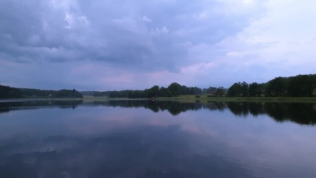 Slow motion. Gorgeous view of sunset on calm summer evening. Lake shore with green trees and plants reflecting in crystal clean mirror water surface. Sky covered with heavy thunder clouds. 