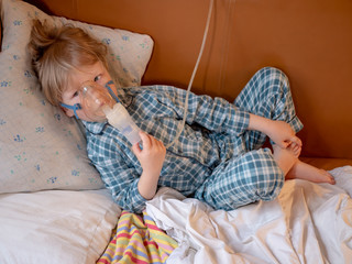 .Funny blond boy in gray pajamas breathes through nebulizer on leather sofa in bedroom. child is...