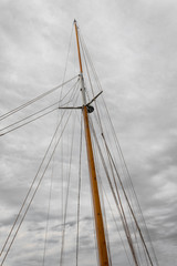 Mast of an old sailing ship with multiple rigging lines against a cold gray sky with clouds, vertical aspect