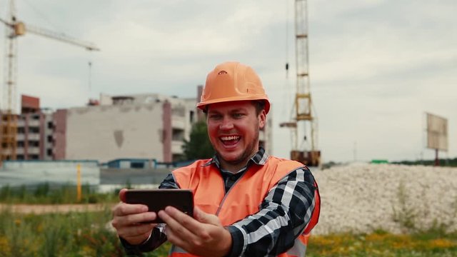 A man in an orange vest and orange helmet takes pictures of himself on a construction site on a mobile phone.