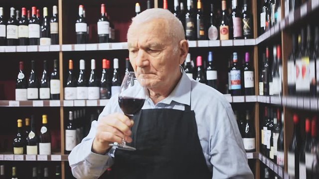 Confident elderly male winemaker holding glass of red wine, checking it in wine store