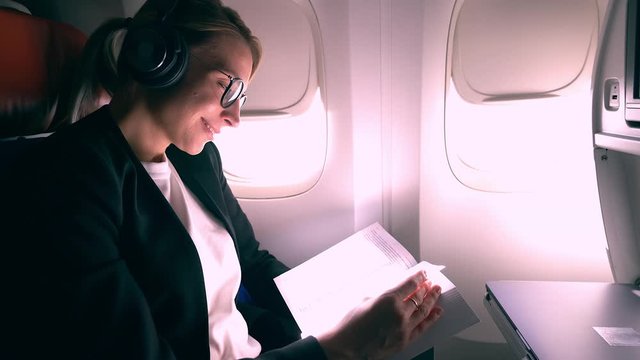 Successful female aircraft passenger in Bluetooth headphones with noise cancellation mode enjoying time for reading literature book best seller and listening favourite music during flight in airplane
