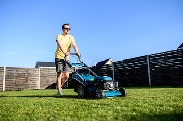 A young man mows a lawn with a lawn mower. Garden works in the backyard, maintenance of the house...