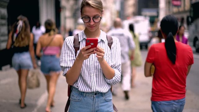 Slow motion effect of attractive caucasian female tourist enjoying communication with friends via mobile application laughing for camera.Millennial hipster girl standing on city street with smartphone