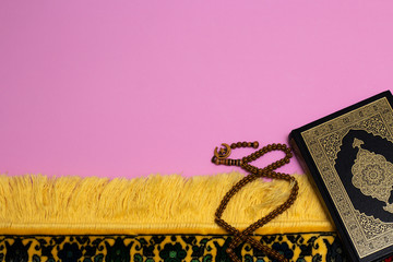 Quran with skullcap and muslim rosary and prayer rug on a pink background