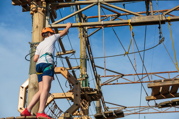 Child in forest adventure park. Kid in orange helmet and white t shirt climbs on high rope trail. Agility skills and climbing outdoor amusement center for children. young boy plays outdoors.