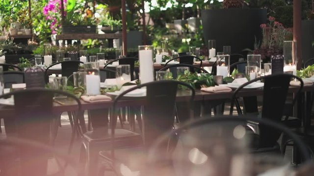 Burning candles on the decorated with green ruscus leaves wedding tables outdoors at the ceremonial veranda, table serving, peopleless tables