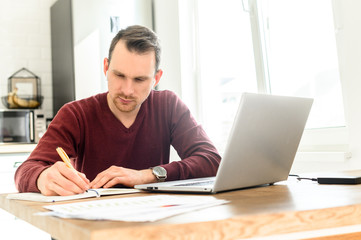 A guy uses laptop for distance study, he sits at the table at home and is writing lecture in a notebook