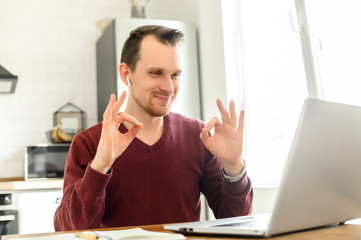 A guy uses laptop for video call, zoom, video communication. He is looking at webcam and shows OK...