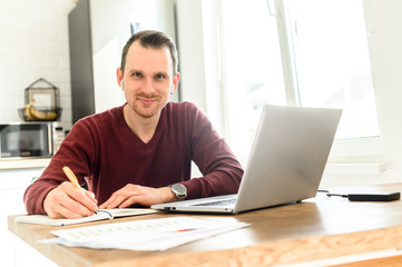 A guy uses laptop for distance study, he sits at the table at home and is writing lecture in a notebook