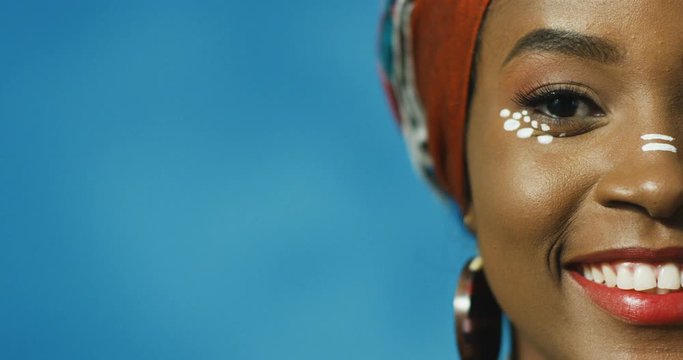 Close up of half face of happy African American beautiful young woman with red lips smiling joyfully to camera on blue wall background. Portrait of pretty female fom Africa.
