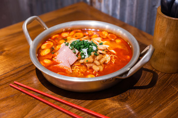 Traditional Korean ramen soup with kimchi, ham, sausage and cheese in silver bowl on wooden table. Korean cuisine. Tasty Asian food.