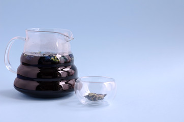 A glass teapot for brewing with blue tea stands on a blue background. Nearby is a small empty bowl. Space for text