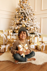 cute little boy with dark hair in cozy clothes posing near decorated Christmas tree with presents