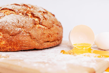 Broken egg on a floured wooden board on a background of appetizing fresh round bread. Homemade baking. Close-up.