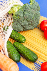 Fresh cucumbers, tomatoes, carrots and broccoli on sprinkled spaghetti in an eco bag. Healthy lifestyle and Mediterranean diet. Delivery during the coronavirus pandemic. Blue background. Vertical.