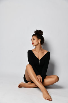 Sensual Brunette Woman In Black Dress Is Sitting Against The Wall With Her Legs Tucked Up Looking Aside