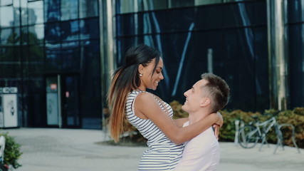 Multiracial family concept: husband spinning around his wife while walking down the street near big office centre one summer day