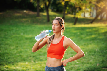 Young gorgeous thirsty slim sportswoman standing with one hand on hip, taking a break after running...