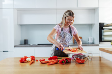 Attractive worthy smiling caucasian blond woman in apron standing in kitchen and putting chopped vegetables into bowl.