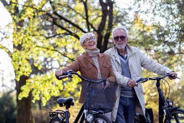 Mature fit couple pushing bicycles in public park talk and smile