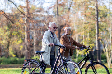 Mature fit couple pushing bicycles in public park talk and smile