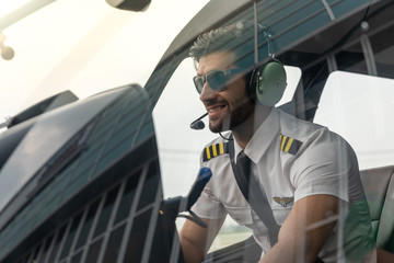 Picture of a male helicopter pilot in his white shirt with tie uniform. He is prepared for his flight. He sits inside a white helicopter with his head phone one ready to take off.