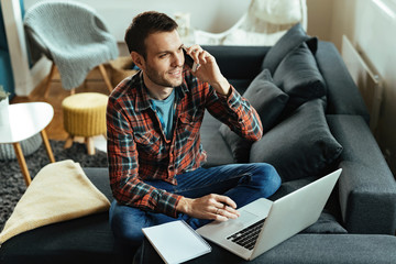 Young man making a phone call while working on laptop at home.