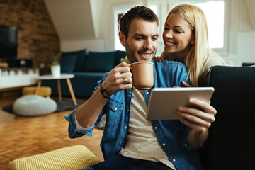 Happy man drinking coffee while using touchpad with his girlfriend at home.