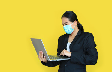 Asian business woman wearing medical shielding mask working with laptop computer isolated on yellow background.