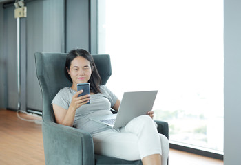 Young asian woman using smartphone and laptop computer working online at home and feel relaxing on armchair.