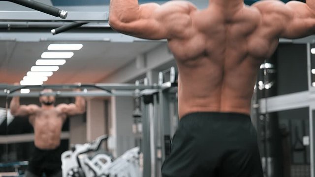 sportsman performing pull-ups on bars during cross-training workout at gym. bodybuilder looks mirror and pulls up on crossbar fitness room bare-torso. rear view athlete's back slow motion.