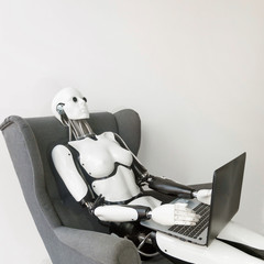 Closeup of white plastic robot working with laptop while sitting in armchair against white wall