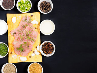 Sliced pieces of meat turkey game filet on a wooden cutting board, garlic, cumin (jeera), salt, fennel, allspice, fenugreek, mustard seeds, oil, green onion, cloves on black wooden background