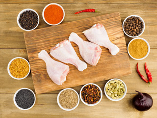 Raw chicken drumsticks on cutting wooden board and spice set: fenugreek, allspice, masala, red chili pepper, red onion, green cardamom, cumin, chili powder, black pepper