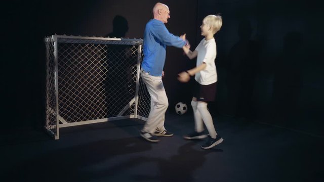 Young Footballer And Old Man Are Playing Soccer Indoors. Boy Is Scoring Goal And Rejoicing