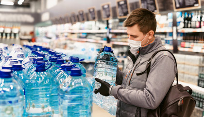 Shopping during the coronavirus Covid-19 pandemic. A young man buys drinking water canisters in a supermarket. Woman in facial mask and gloves to prevent infection.